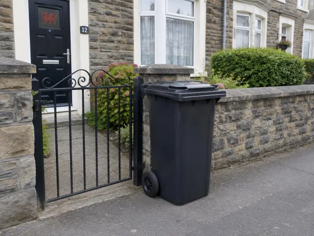 A black wheelie bin with its lid shut by a front garden gate